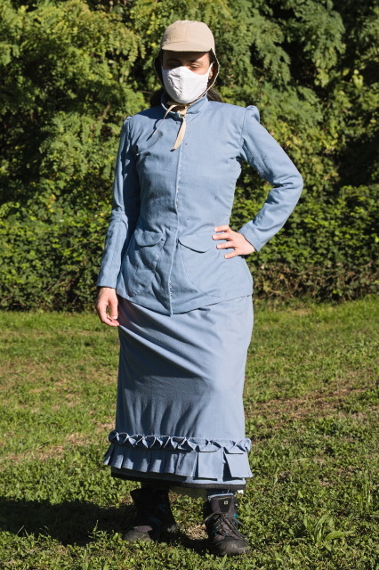 A woman wearing a light blue linen-cotton outfit with an ankle length skirt with box pleats at the hem and a fitting jacket with a high collar and big applied pockets. Proper (almost) hiking boots (modern) complete the (historical) hiking dress, while the non-matching headgear and face mask interfere with the collar of the jacket (uooops).