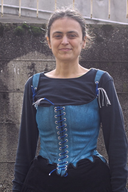 the top half of a woman wearing a garment in the shape of 18th century stays, but unboned and relatively soft, in sparkling blue, badly spiral laced closed with a length of paracord