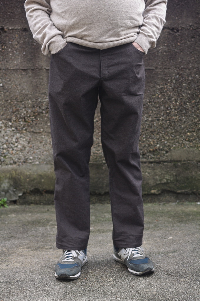 The lower torso and legs of a man wearing a pair of jeans-like trousers in brown fabric; he's keeping his hands in the pockets.