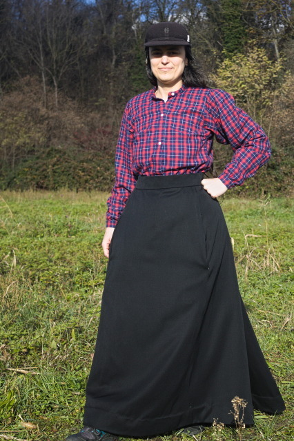 a woman walking on grass with a 1880s plaid shirt, an almost floor length 1890s black wool skirt and a glimpse of a 2020s hiking boot