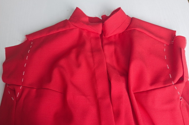 the top half of a shirt in red wool: there are basting lines where the sleeves will attach, and the collar is attached, but still not hemstitched closed, the front placked is finished, but still has no buttonholes / buttons.