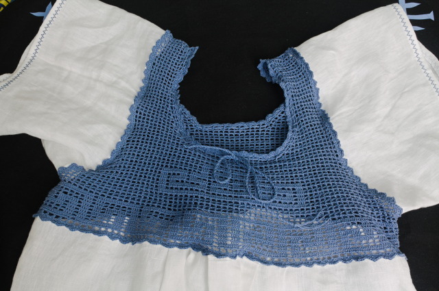 The top half of a nightgown with a yoke in filet crochet with a meander pattern in light blue yarn, and rectangular body and sleeves in white linen fabric. The hem on the sleeves is sewn with a decorative stitch in light blue thread.