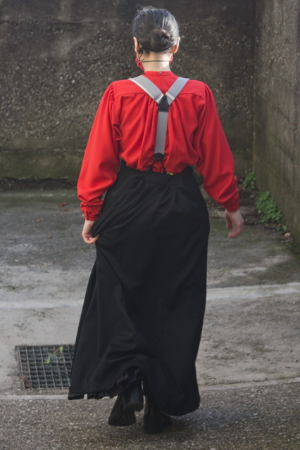a woman walking away from the camera wearing a black long skirt, grey braces, and a red shirt with wide sleeves and a lot of fabric gathered in the middle part of the fitted yoke. The collar is just a collar band, and there is a button in the nape of the neck for a detached collar that is not being worn.