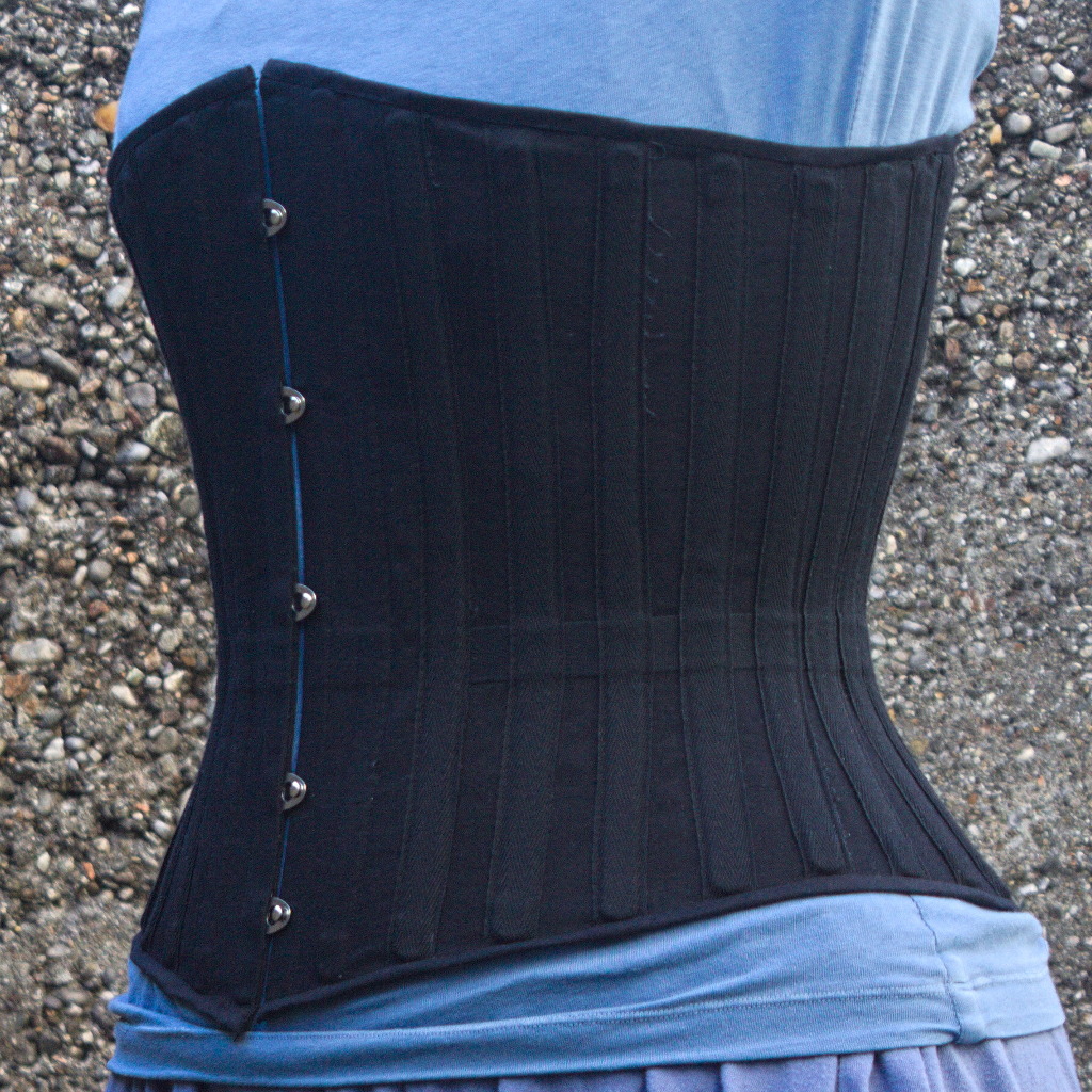 a black coutil midbust corset, from a 3/4 front view, showing the busk closure, a waist tape and external boning channels made of the same twill tape and placed about 1-2 cm from each other at waist level.