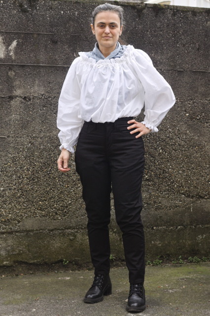 a woman wearing black jeans and a very full white voile shirt with a wide neck; both the neck and the cuffs are gathered with elastic and have a ruffle on top. there is a light blue scarf to fill the neckhole.