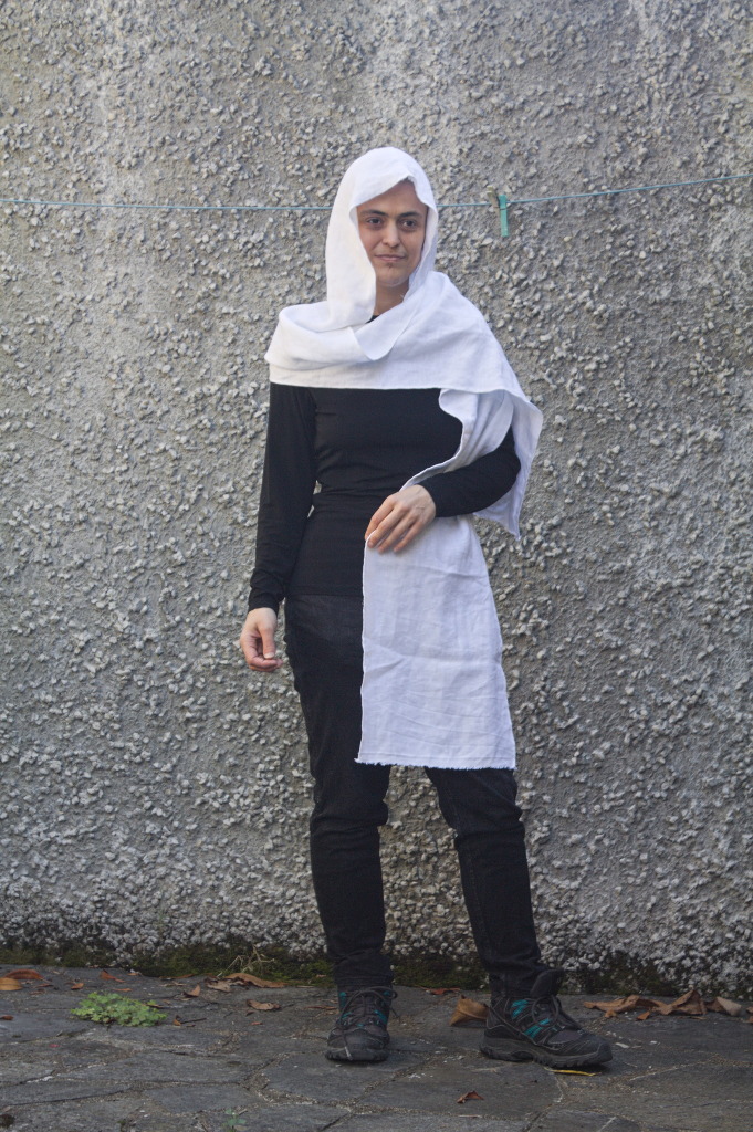 A woman wearing some sort of white veil that covers the head and has two scarf-like appendages, one wrapped around the shoulders and falling on the back, and one falling on the front side, kept in place with one arm.