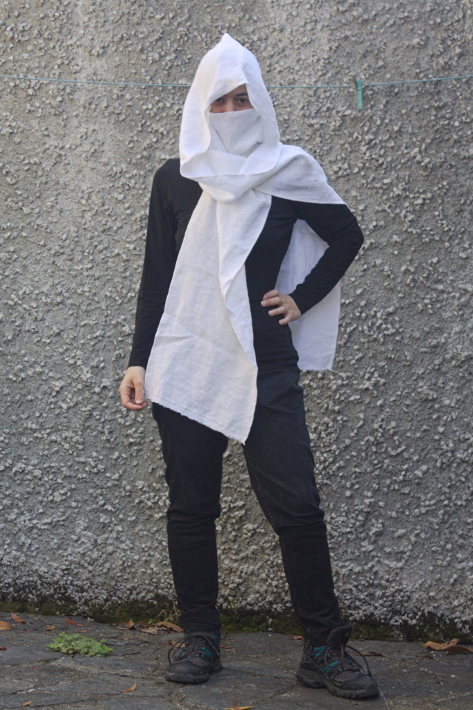 A woman wearing what looks like a deep hood over some sort of fabric face mask, showing only the eyes, in a way that resembles characters in a video game. There are again two scarf-like edges that fall on the front and back.