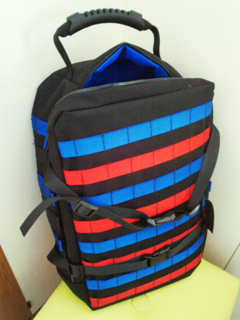 picture of a mostly parallelepipedal backpack in black fabric with blue and red webbing stripes on the front and black webbing compression straps. the lining can be seen at the top and it's light blue.
