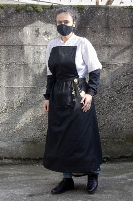 A person dressed in late 1800 clothing with a black apron with a gathered front and triangular side panels that wrap to the sides of the shirt, plus a bib pinned up on the shirt, and black sleeve protectors. There is a chatelaine pinned at the waist with an Ada Lovelace pin, with fake punched cards, a pencil and steampunkish pendants. Also, the shoes are horribly wrong, and we don't talk about the leggings that can be seen at the ankles.