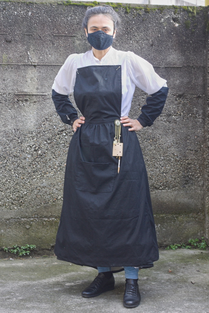 A person dressed in mostly late 1800 clothing with a black apron with a gathered front and triangular side panels that wrap to the sides of the shirt, plus a bib pinned up on the shirt, and black sleeve protectors. There is a chatelaine pinned at the waist with an Ada Lovelace pin, with fake punched cards, a pencil and steampunkish pendants.