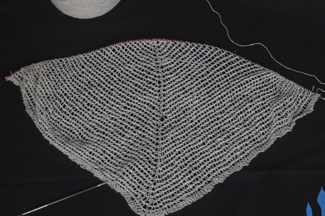 a work in progress triangular white shawl in white thin and lightly fuzzy yarn, with a very simple lace pattern that consists in rows of holes parallel to the two borders.