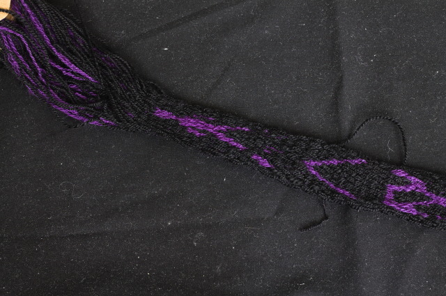 an in-progress tablet woven band in black with purple line based designs; the one closest to the tablets is a stick figure with a few missing bits