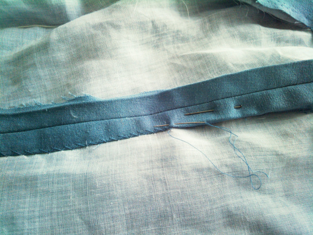 The inside of a seam in a garment made of light blue cloth lined in white; the raw edges have been pressed open, a fold has been pressed in them and one side is being sewn down to the lining with herrinbone stitches.