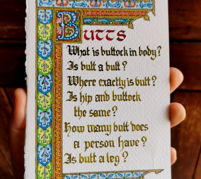 A closer crop of my hand holding paper, to show the colours of the red and green inks used for the text.

The word 'Butts' is calligraphied at the top in red ink uncial font; the 'B' is illustrated inside its own square.

Below, in green ink, blackletter font;

"What is buttock in body?
Is butt a butt?
Where exactly is butt?
Is hip and buttock the same?
How many butt does a person have?
Is butt a leg?"

The text is framed above, left, and below with an illustrated border of leaves and repeating patterns in reds, browns, blues, and greens, with gold highlights.