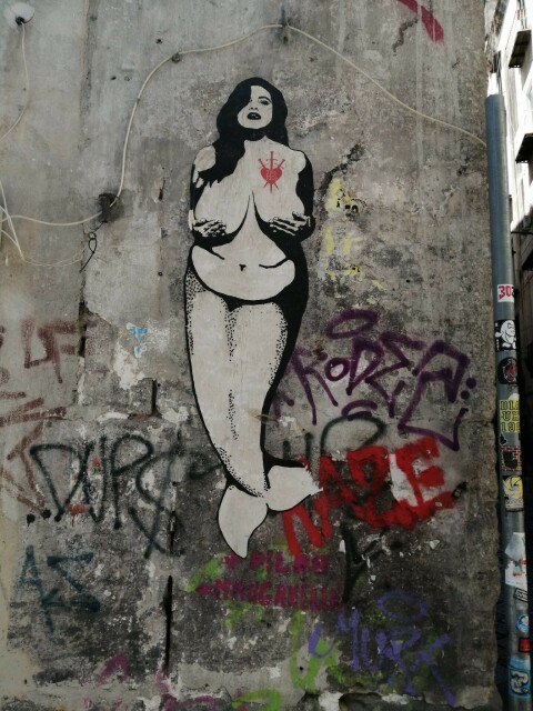 A piece of street art (stencil style) of a curvy mermaid with long dark hair and a red tatoo-like symbol on the right side of her chest that looks like a heart pierced by three swords
