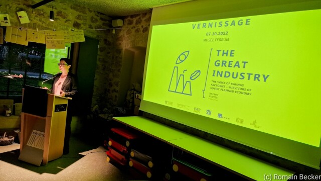 A speaker in front of a screen. The screen shows the title of the exhibition.