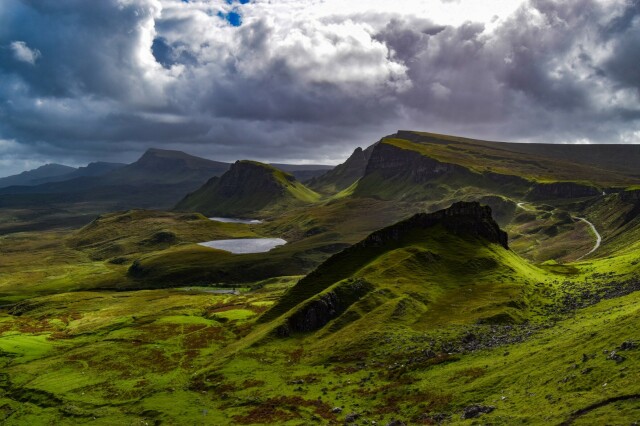 A view of the Quiraing mountain range on the Isle of Skye, Scotland. The picture is a mix of sharp cliffs and soft hills, covered in vibrant green vegetation. Two small lakes are nestled amongst the hills. The sky is cloudy which casts shadows on the ground. There are no people on the picture. Sheep can be seen as little white dots on the lower part of the picture.