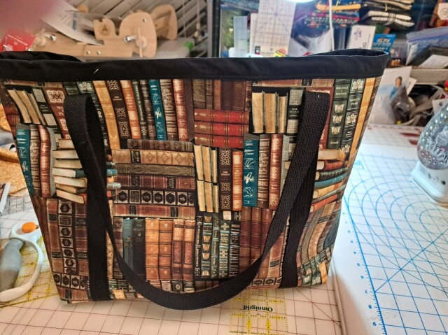 A tote bag made of book-themed fabric, with heavy-duty long black handles.