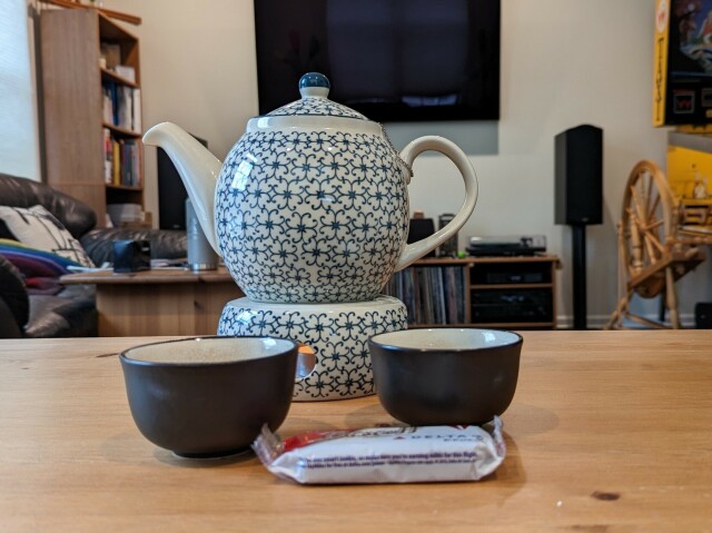 A blue and white teapot sits on a matching base with a tea light underneath. Two cups are set out in front of the teapot, along with a Delta Airlines packet of Biscoff cookies. 