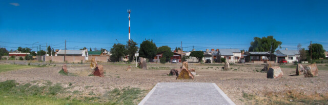 A group of large stones arranged in a circle on a gravel surface, itself surrounded by grass. A paved path leads to the circle, blue sky and buildings behind. Plaza de Colonnos, Gaiman, Argentina : a Gordsedd Circle. Semudobia on Wikimedia Commons.