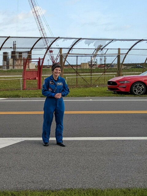 Outdoor day scene of a paved road, at a side of which is a metallic net fence. On the road stands a woman in her mid forties with short black hair, in a blue pilot flight suit. Close to the woman a red convertible car can be partially seen.