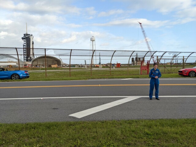 Outdoor day scene of a paved road, at a side of which is a metallic net fence. On the road stands a woman in her mid forties with short black hair, in a blue pilot flight suit. Beyond the fence is a white rocket on a launch pad that has a black service tower next to it. To the right of the pad are a white water tank tower and a crane. Close to the woman a red convertible car can be partially seen. A similar blue car can be partially seen on the left.