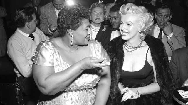 Black and white photo of Marilyn and Ella seated and looking glamourous. Ella is speaking to Marilyn and smiling and she is smiling and listening.
