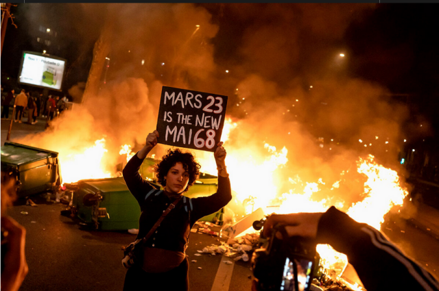 A person at night looking determined, holding a sign saying "Mars 23 is the new Mai 68" in front of a garbage fire, pretty much summarizing everything already...