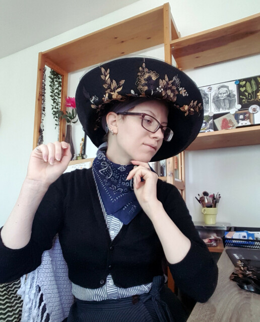 A selfie photo of Sini wearing comfy normal clothes, a cardigan, bandana and striped shirt, with a very large very different vibe black hat on her head. Its brim has lace netting embroidered with gold, purple, black and silver flowers pinned to it. It looks rather loud and embellished for modern sensibilities. She is looking down with a gently pleased and playful expression on her face. There are postcards, make up brushes and a decorative vase with a pink plastic flower in it in the background.