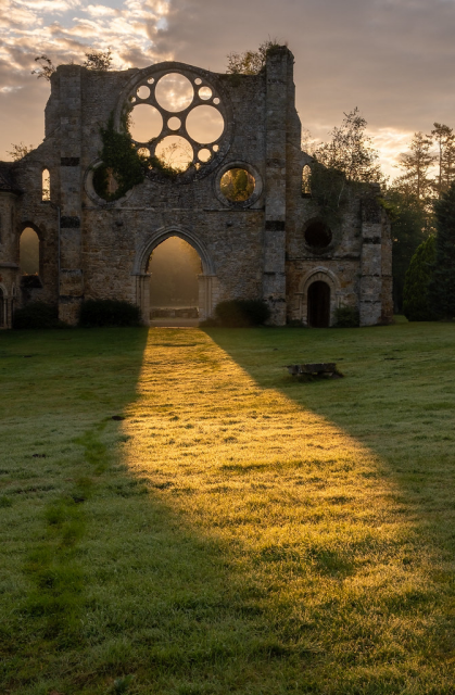 In this striking photo by Andrew Hayman, we see the long, slanting light of the setting sun pour through the door of a ruined abbey church in the Île-de-France. Above the door is the intact stone framework of the abbey’s east rose window, through which we see the evening sky and clouds. 