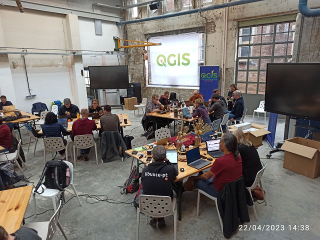 A big group of QGIS developers sitting around multiple sets of tables in an industrial meeting space, decorated with QGIS signs, discussing and pogramming.