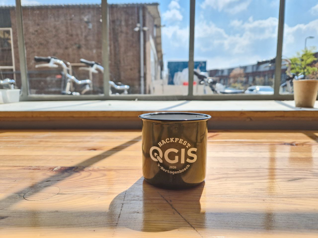 A camping mug with the QGIS Hackfest logo, dating back to 2020 on a table in front of a window with the sun shining through the window