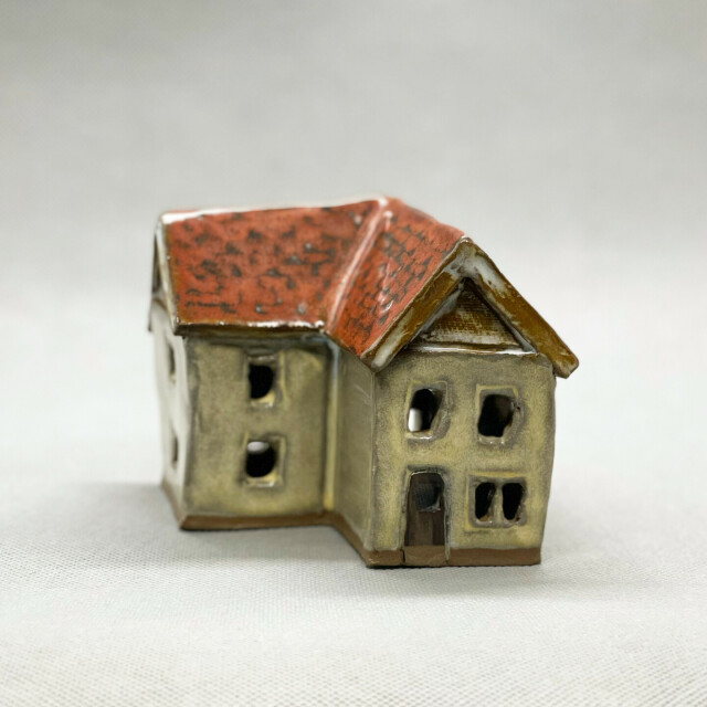 A small ceramic house. It has yellow walls and a red roof. Windows and doors are cut into the walls. The house has an L-shaped layout and is two storeys tall with a peaked roof. 