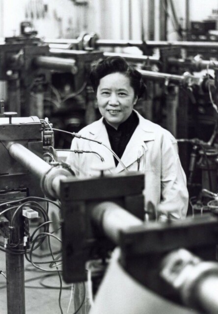Chien-Shiung Wu standing behind the beam line of her apparatus. She is wearing a white lab coat and smiling at the camera.