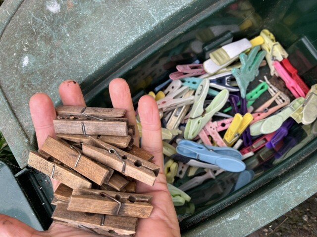 A photo of a hand holding wooden clothes pegs, above a box of plastic begs.