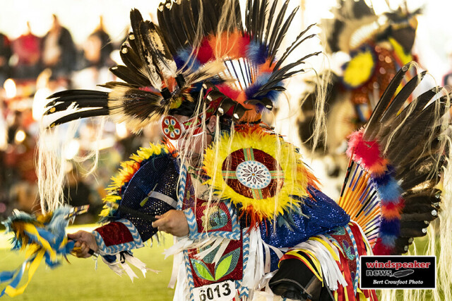 A Fancy Dancer is dressed in colourful regalia with feathers and beadwork adornment. He is dancing in a grassy area in the powwow arbour.