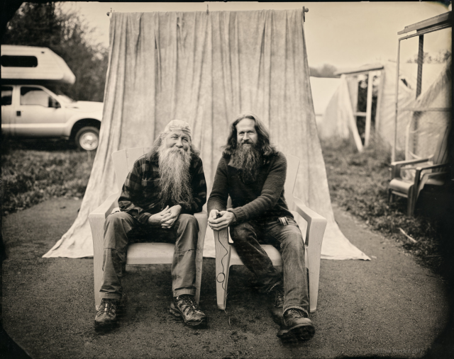 Two men in their 60s with long hair and long beards sit side by side, facing the camera while leaning toward each other. A photographic backdrop curtain hangs behind them, but they're clearly outside on a driveway. To the left, the front end of a pickup truck with an over-the-cab camper is visible. On the right are the front entry ways to a series of greenhouses.  The image itself was made using a glass negative technique from the 1800s. Not quite black and white , it's more of a light tan and black image. We've been together for twenty-five years, married for the last seven.