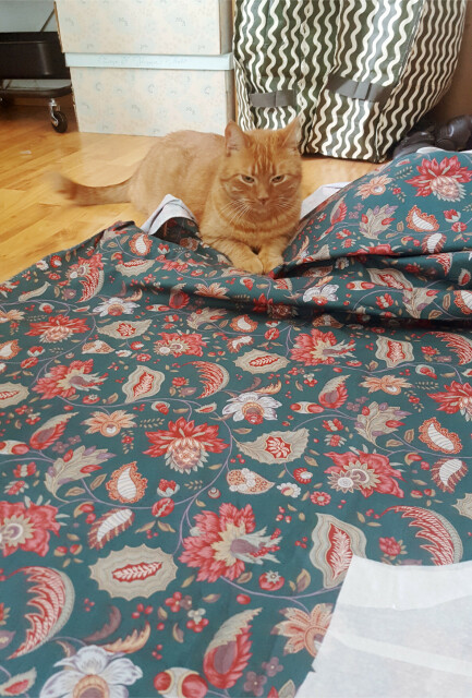 A photo of a ginger tabby cat resting on a rather loud flower pattern fabric that has been laid out on the floor to cut it. The base colour is a petrol blue, but the red and light green flowers are the much more prominent element. The cat seems determined but also like he knows he's not supposed to be where he is.