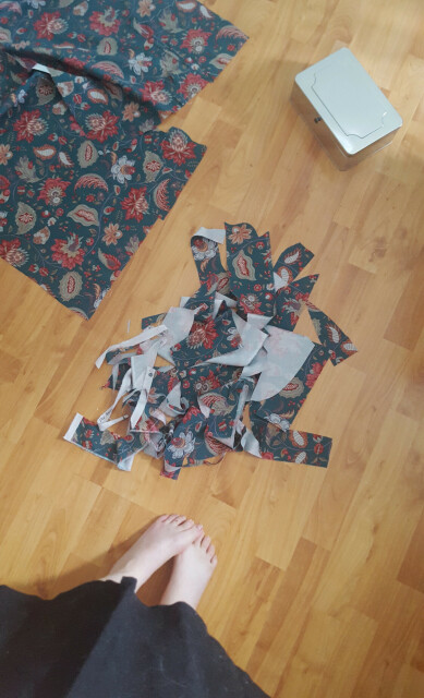 A photo looking down on a rather small pile of extremely small fabric scraps. It doesn't look like it could be feasibly used even for quilting, the pieces are such slim slivers. A bigger length of the already cut skirt pieces is in the frame, as well as Sini's toes and a sewing supplies tin.