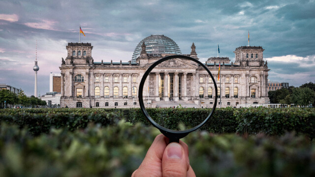Image of the Bundestag with a hand holding a magnifying glass on the foreground