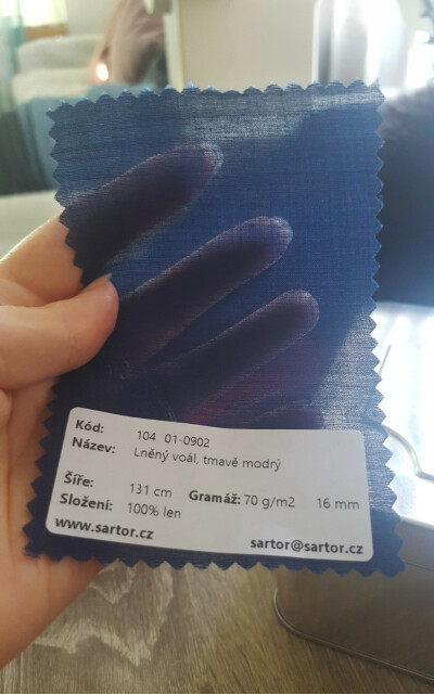 A photo of a disembodied hand holding up a fabric swatch or sample. It is roughly the size of a palm, cut with pinked edges. The fabric is see through seer, an indigo dark blue, and densely woven from very fine linen thread with a reasonable amount of natural variation in the thread weight. It is preposterously fine for a linen fabric. There is a sticker with the fabric information, written in Czech. Roughly translated it declares the item number, that it is linen voile, dark blue, width of 131cm, 100% linen, and of the weight 70g per square metre. Which is also preposterously light for linen. There is an url on the sticker for Sartor Bohemia, www.sartor.cz