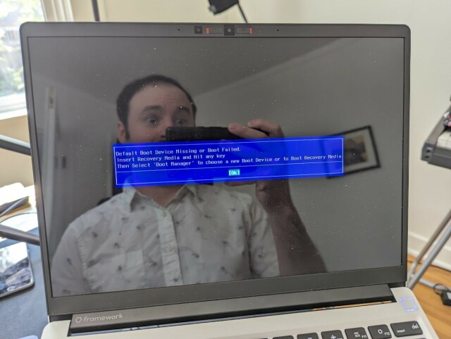 A laptop with a blue BIOS screen reading:

Default Boot Device Missing or Boot Failed

The photographer is making a silly surprised face in the screen reflection.