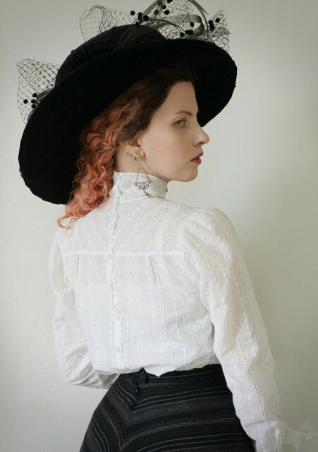 A photo portrait of Sini wearing a very large black hat, a black skirt and a very dainty white blouse with white embroidery on it. The view is from the back, with her face turned to show her profile, cropped to upper hip and upward. The blouse buttons from the back, with the upper back yoke cut on the bias and having very closely spaced white pearl buttons, the lower back with them wider apart. The fabric is so sheer you can see the very narrow seams and a corset cover straps beneath it. The blouse blouses down from the yoke and is cinched in by the skirt waist.