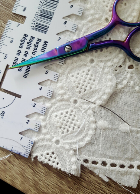 A very very close up photo of the collar in progress, with a repeat of the embroidered pattern being sewn on another piece, going crosswise. The embroidered bit has a continuing line of embroidery that surrounds a chained circle motif, and this is in the process of being sewn down by hand with a tiny needle, with an absolutely tiny seam allowance. The stitches themselves are nearly invisible, the white thread getting lost in the white embroidery. There is a seam gauge and scissors for scale. The scale is minuscule.