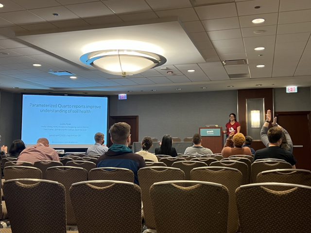 View from the audience of a woman presenting at posit::conf(2023) with the slide title of Parameterized Quarto Reports Improve Understanding of Soil Health. 