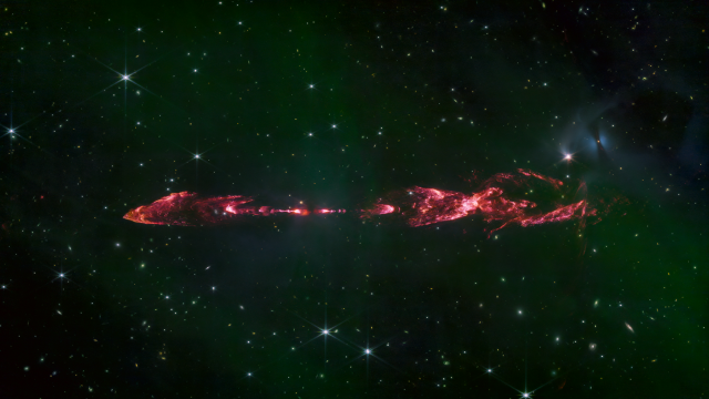 A purple-red jet of emission with a series of knots and bowshocks, symmetric about an invisible protostar, seen against as field of stars and galaxies, with green nebula emission across the image.