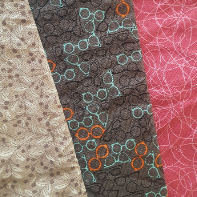 A pleasantly square shaped photo of three different print fabrics fanned out on top of each other, showing a sliver of each. Starting from the left they are a beige fabric with delicate brown and paler tan berry plants, reminiscent of Nordic blueberries. The next one is a gray base with black, teal and orange eyeglasses, both vintage and hipster shapes, haphazardly arrayed to fill the entire space. The third one is a nice cold tone berry red or pink with lighter pink abstract squiggles on it. Side by side they vaguely look like they could all be worn by the same person without immediately being sent to fashion jail.