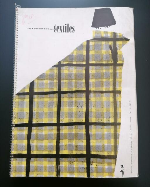 Copertina di un catalogo di moda internazionale, stampato in Olanda nel 1957, raffigura una donna avvolta in un largo mantello tartan giallo, grigio e nero, da cui emergono solo gli occhi, con un cappello nero sulla testa, e la scritta in nero International textiles.