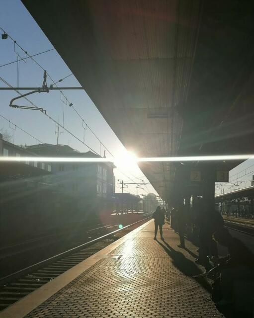 Pendolari, solo ombre nel sole che si alza oltre i binari, aspettano rassegnati il treno, in ritardo.