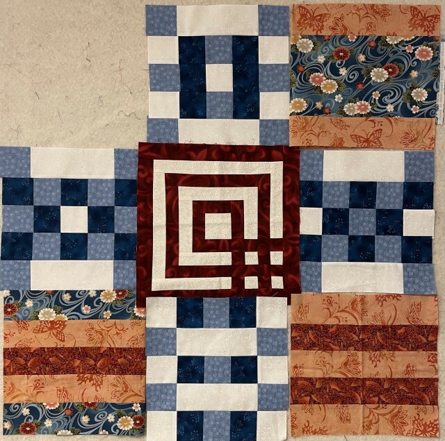 A nine patch quilt missing the top left square. In the center is a red and white square that looks like the bottom right quarter is a grid of red and white lines. The rest of that block is just alternating red and white, with red on the outside. 

At the top and bottom and two sides are blue, dark blue, and white pieced squares. None of them is exactly like the other, but mostly the squares of color alternate.

At the bottom left is a square with a blue Japanese floral print, orange batik, and dark orange batik. On the bottom right is a square of just alternating lighter orange and darker orange batik. On the top right is a square with the lighter orange batik on top and bottom and the blue Japanese floral in the middle.